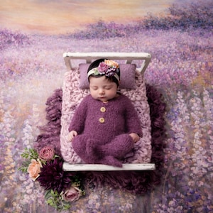 Puede incluir: Un bebé recién nacido con un conjunto de punto morado y una diadema floral, acostado en una cama blanca en miniatura con una manta y almohada moradas. El fondo es un campo de lavanda pintado.