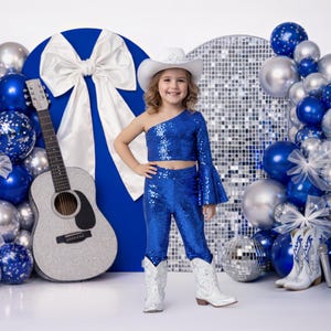 Może przedstawiać: Młoda osoba w niebieskim cekinowym stroju, białych kowbojskich butach i białym kapeluszu kowbojskim stoi przed niebiesko-srebrnym łukiem z balonów. Srebrna gitara i kule dyskotekowe również znajdują się na scenie.
