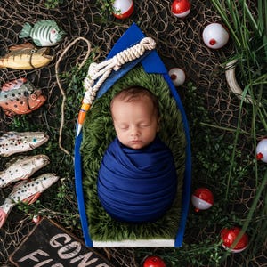 May include: A newborn baby wrapped in a blue blanket, nestled in a blue boat-shaped prop with green faux grass. The scene is decorated with fishing bobbers, wooden fish, and a sign that reads "Gone Fishin'."