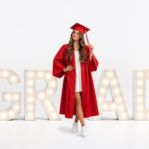 May include: A person in a red graduation gown and cap stands in front of illuminated letters spelling "GRAD". The person is wearing a white dress and white sneakers. The graduation cap has a red tassel.