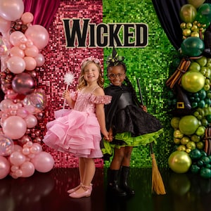 Op de afbeelding: Twee jonge meisjes in kostuums poseren voor een achtergrond met pailletten met het woord "WICKED". Eén draagt een roze jurk en houdt een toverstaf vast, de ander een heksenkostuum met een bezem. Roze en groene ballonnen versieren de zijkanten.