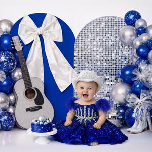 Może przedstawiać: Dziecko w niebieskiej sukience z cekinami i białym kapeluszu kowbojskim uśmiecha się na tle z niebieskimi i srebrnymi balonami, gitarą i ciastem. Tło zawiera dużą białą kokardę i kulę dyskotekową.