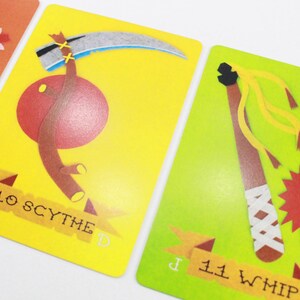 Puede incluir: Dos cartas de juego ilustradas con fondos amarillo y verde. La carta de la izquierda muestra una guada&ntilde;a roja y marr&oacute;n con el texto "10 SCYTHE". La carta de la derecha muestra un l&aacute;tigo marr&oacute;n con un borla amarillo y rojo y el texto "11 WHIP".