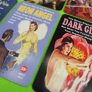 May include: A collection of vintage-style cards with retro illustrations and text. One card features a woman in a yellow dress with the text "NEON ANGEL." Another card displays a woman with a flower and the text "DARK GIFT."