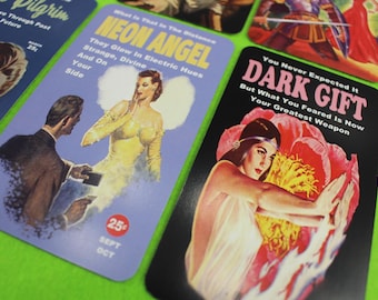 Pode incluir: Uma coleção de cartões em estilo vintage com ilustrações e texto retrô. Um cartão apresenta uma mulher em um vestido amarelo com o texto "NEON ANGEL". Outro cartão mostra uma mulher com uma flor e o texto "DARK GIFT".