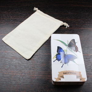 Op de afbeelding: Een tarotkaartendeck met een witte achtergrond en een blauwe en zwarte vlinder op de voorkant. De kaart is getiteld "Twee van Staven". Het deck wordt geleverd met een witte stoffen tas.