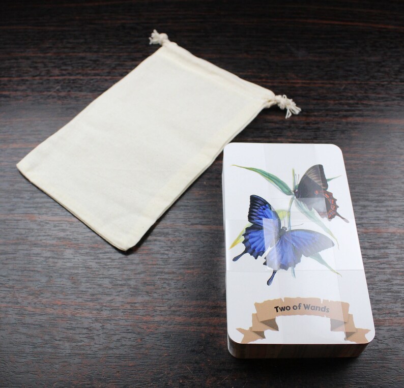 Op de afbeelding: Een tarotkaartendeck met een witte achtergrond en een blauwe en zwarte vlinder op de voorkant. De kaart is getiteld "Twee van Staven". Het deck wordt geleverd met een witte stoffen tas.