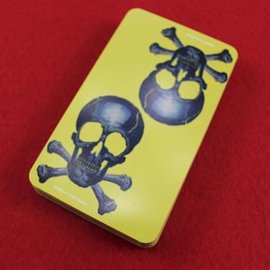 May include: A deck of tarot cards with a yellow background and two illustrations of skulls with crossbones. The skulls are dark blue with yellow eye sockets. The text "Buddy's Tarot Decks" is printed on the bottom.