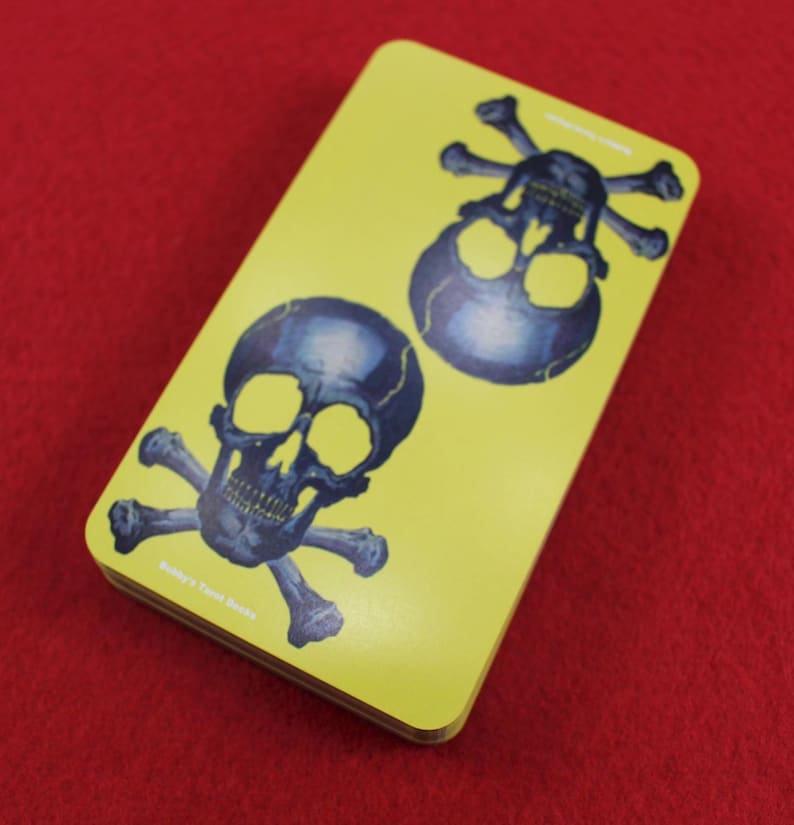 May include: A deck of tarot cards with a yellow background and two illustrations of skulls with crossbones. The skulls are dark blue with yellow eye sockets. The text "Buddy's Tarot Decks" is printed on the bottom.