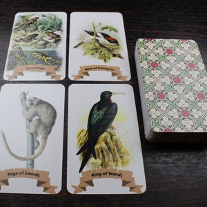 Op de afbeelding: Een set tarotkaarten met illustraties van dieren en natuur. De kaarten zijn in een rasterpatroon op een houten oppervlak gerangschikt. De kaarten omvatten de Tien van Bekers, de Negen van Pentakels, de Page van Zwaarden, de Koning van Staven en een kaartspel met een groen-rood patroon.