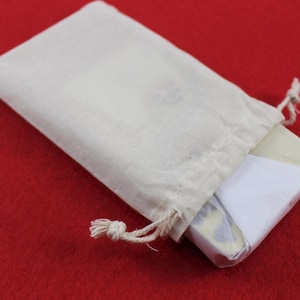 May include: A small, off-white drawstring bag with a rectangular item partially visible inside. The item is wrapped in white and patterned paper. The bag is on a red surface.