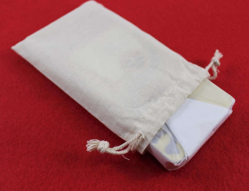 May include: A small, off-white drawstring bag with a rectangular item partially visible inside. The item is wrapped in white and patterned paper. The bag is on a red surface.