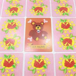 Puede incluir: Una carta de juego rosa con un dise&ntilde;o de estrellas amarillas y una rosa roja. La carta presenta un oso marr&oacute;n de dibujos animados con una calavera y huesos cruzados en el pecho. La carta dice "13 BEAR C".