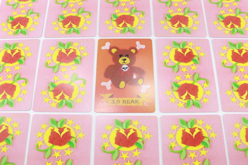 Puede incluir: Una carta de juego rosa con un dise&ntilde;o de estrellas amarillas y una rosa roja. La carta presenta un oso marr&oacute;n de dibujos animados con una calavera y huesos cruzados en el pecho. La carta dice "13 BEAR C".