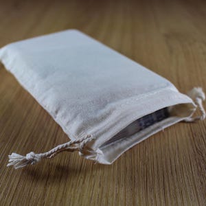 Puede incluir: Una peque&ntilde;a bolsa rectangular de lona blanquecina con cierre de cord&oacute;n. La bolsa est&aacute; sobre una superficie de madera. La bolsa est&aacute; ligeramente abierta, revelando una vista del contenido.