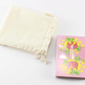 Puede incluir: Una baraja de cartas rosa con un dise&ntilde;o floral rojo y amarillo. Las cartas est&aacute;n en una bolsa de tela blanca con cord&oacute;n.