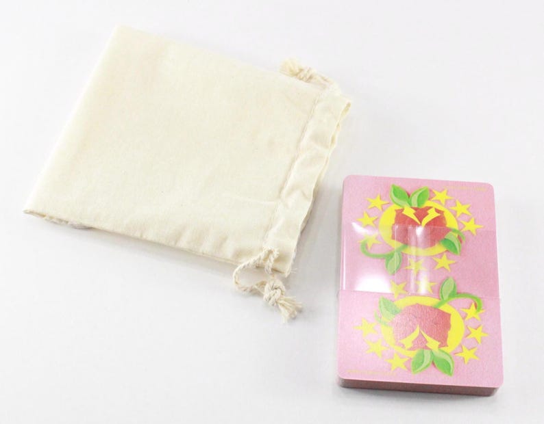 Puede incluir: Una baraja de cartas rosa con un dise&ntilde;o floral rojo y amarillo. Las cartas est&aacute;n en una bolsa de tela blanca con cord&oacute;n.