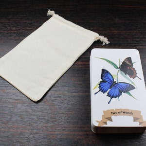 Op de afbeelding: Een witte stoffen tas met een trekkoord ligt naast een tarotkaart met een afbeelding van een blauwe en zwarte vlinder. De kaart toont "Twee van Staven".
