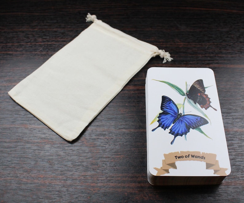 Op de afbeelding: Een witte stoffen tas met een trekkoord ligt naast een tarotkaart met een afbeelding van een blauwe en zwarte vlinder. De kaart toont "Twee van Staven".