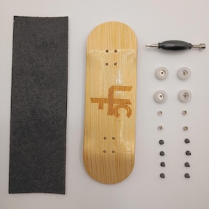 May include: A fingerboard deck with a light wood grain finish and a brown logo featuring the letters LFC, accompanied by a rectangular sheet of black grip tape, one black truck, four white wheels, and a set of small metal hardware components.