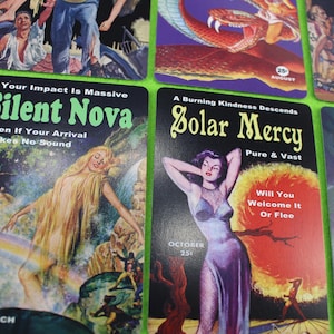 Könnte beinhalten: Eine Sammlung von Kunstdrucken im Vintage-Stil mit Retro-Illustrationen und Text. Die Drucke zeigen lebendige Farben und Bilder, darunter eine Frau, eine Schlange und Texte mit der Aufschrift "Silent Nova" und "Solar Mercy".