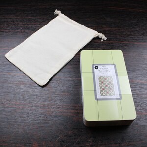 Op de afbeelding: Een groene en witte quiltliniaal met een rasterpatroon en een klein kaartje met een quiltblokdesign. De liniaal is verpakt in een witte trekkoordtas.