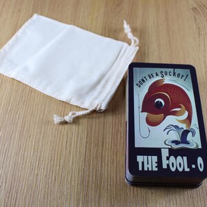 Puede incluir: Una baraja de cartas con un dise&ntilde;o vintage que presenta un pez rojo en un anzuelo, el texto "DON'T BE A SUCKER!" y "THE FOOL-O". Una peque&ntilde;a bolsa con cord&oacute;n de color blanquecino est&aacute; junto a la baraja sobre una superficie de madera.