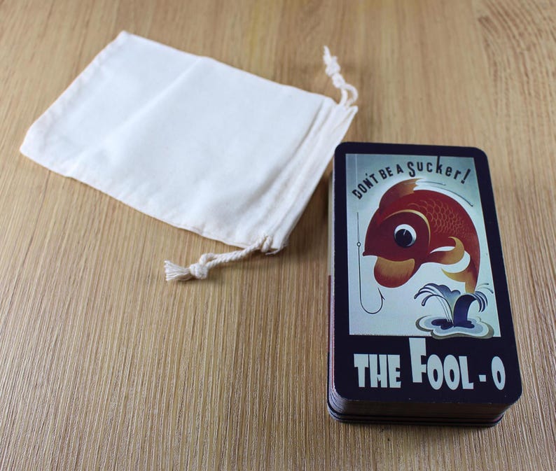 Puede incluir: Una baraja de cartas con un dise&ntilde;o vintage que presenta un pez rojo en un anzuelo, el texto "DON'T BE A SUCKER!" y "THE FOOL-O". Una peque&ntilde;a bolsa con cord&oacute;n de color blanquecino est&aacute; junto a la baraja sobre una superficie de madera.