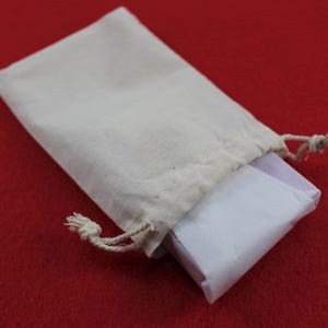 Pode incluir: Uma pequena bolsa com cord&atilde;o de cor creme com um pacote embrulhado em papel branco. A bolsa &eacute; feita de um tecido de cor natural e repousa sobre uma superf&iacute;cie vermelha. O cord&atilde;o &eacute; amarrado no topo.