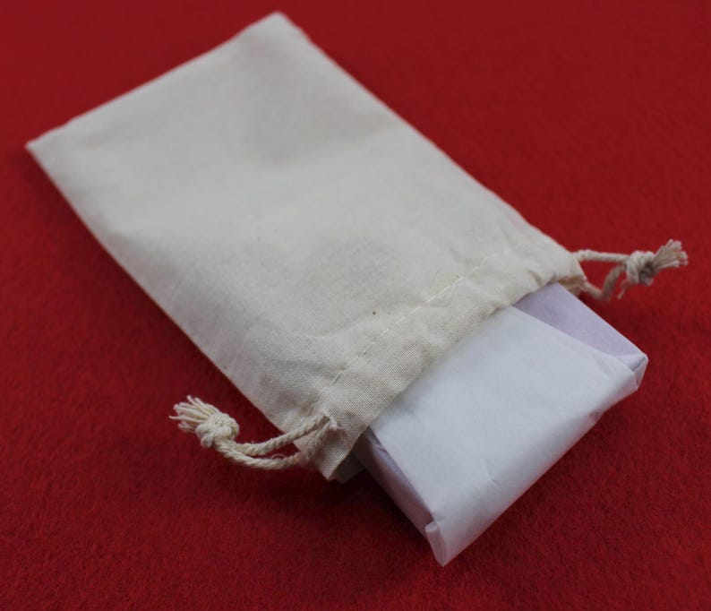 Pode incluir: Uma pequena bolsa com cord&atilde;o de cor creme com um pacote embrulhado em papel branco. A bolsa &eacute; feita de um tecido de cor natural e repousa sobre uma superf&iacute;cie vermelha. O cord&atilde;o &eacute; amarrado no topo.