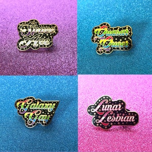 May include: Eight enamel pins with a space theme and LGBTQ+ identities. The pins are black with gold outlines and feature rainbow, bisexual, lesbian, transgender, pansexual, non-binary, and asexual pride flags. The pins read "Space Ace", "Quasar Queer", "Galaxy Gay", "Lunar Lesbian", "Nebula Nonbinary", "Planetary Pan", "Twilight Trans", and "Blackhole Bi".