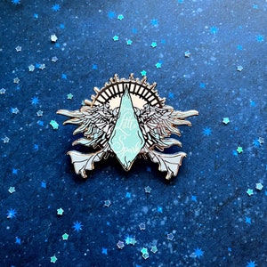 May include: A silver enamel pin with a blue and white crystal design. The crystal has the words "Your Little Spark" written on it. The crystal is surrounded by two wings and a circle with a sun design.
