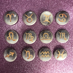 Puede incluir: Una colección de doce botones redondos con signos del zodiaco. Cada botón presenta un símbolo del zodiaco dorado sobre un fondo gris y blanco con efecto de estrellas. Los botones están dispuestos en tres filas de cuatro.