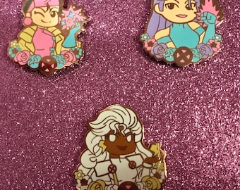 May include: Three enamel pins featuring cartoon characters with floral accents. The first pin features a pink and yellow character with a pink star. The second pin features a purple character with a blue and pink star. The third pin features a white character with a yellow star.
