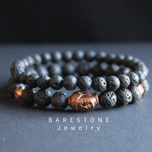 Puede incluir: Dos pulseras de piedra de lava negra con un dije de cabeza de Buda de cobre. Las pulseras están apiladas una encima de la otra. BARESTONE Jewelry está impreso debajo de las pulseras.