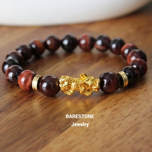 May include: A red tiger's eye and red jasper beaded bracelet with a gold dragon charm. The bracelet is on a wooden surface. BARESTONE Jewelry.
