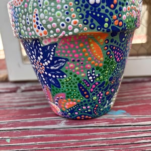 May include: A hand-painted flower pot with a vibrant, colorful design. The pot features a base color of green, adorned with blue, pink, orange, and white dot patterns and floral motifs. The rim has a lighter green hue, complementing the overall design.