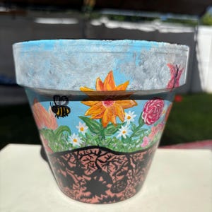 May include: Hand-painted ceramic flower pot with a blue sky and cloud design at the top. The pot features a vibrant floral scene with a bee, flowers, and a butterfly. The bottom has a black and pink decorative pattern.