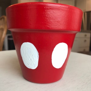 May include: A red painted flower pot with two large white ovals on the front. The pot is made of terracotta and has a smooth, glossy finish. The design is reminiscent of a famous cartoon character.