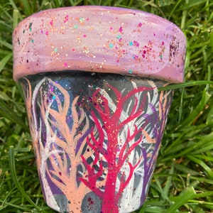 May include: A hand-painted flower pot with a forest design. The pot features a black base with colorful tree illustrations in shades of pink, white, and purple. The rim is painted light pink and covered in glitter.