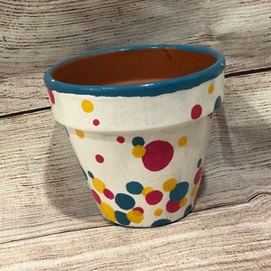 May include: A white terracotta flower pot with a blue rim and colorful polka dots in red, yellow, and blue.