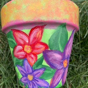 May include: A terracotta flower pot painted with green, red, purple, and yellow flowers. The pot is decorated with a green, yellow, and pink gradient on the top rim.