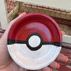 May include: A hand-painted, round, red, white, and black ceramic dish. The dish is designed to resemble a Poké Ball, with a red top, white bottom, and a black stripe and circle in the center. The dish is held in a person's hand.