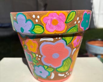 May include: A hand-painted terracotta flower pot with a brown base and rim. The pot is decorated with large, colorful flowers in shades of pink, purple, yellow, and teal, with green leaves. White dots accent the design.