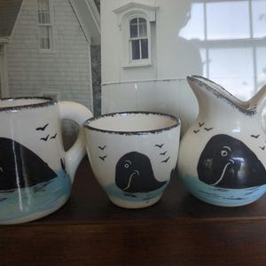 May include: A set of three white ceramic mugs with black whale illustrations and blue water accents. The mugs are decorated with small black birds.