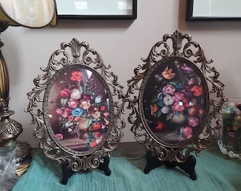 May include: Two ornate, oval-shaped picture frames with floral still life prints. The frames have a gold finish and are supported by black stands. The floral arrangements feature a variety of colorful blooms, including red, pink, and blue flowers.