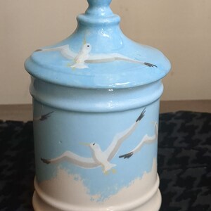 May include: A white ceramic jar with a blue lid and a blue and white seagull design. The jar has a white base and a blue top with a white knob.