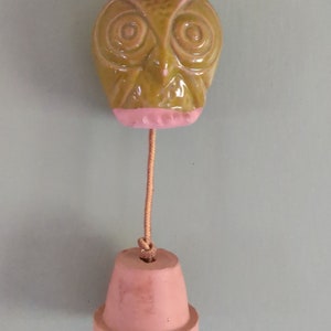 May include: A ceramic owl with pink accents hangs from a brown cord with a terracotta pot at the bottom.