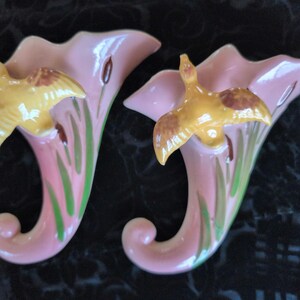 May include: Pair of vintage wall vases in a soft pink hue, each featuring a yellow bird in flight. The vases have a curved, trumpet-like shape with green and brown accents, creating a charming, nature-inspired design.