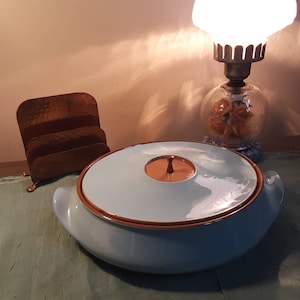 May include: A light blue ceramic casserole dish with a brown rim and a matching lid. The lid has a small knob on top. The dish is sitting on a green surface.